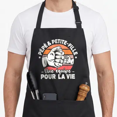 Tablier de cuisine noir avec illustration d'une poignée de main entre une grande et une petite main sur fond dégradé orange et rouge.