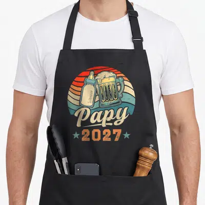 Tablier de cuisine noir avec illustration vintage montrant un biberon et une bière sous l'inscription Papy 2027.