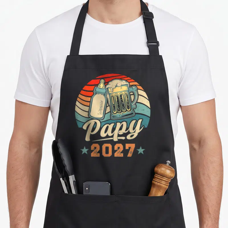 Tablier de cuisine noir avec illustration vintage montrant un biberon et une bière sous l'inscription Papy 2027.