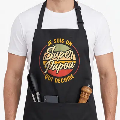 Tablier de cuisine noir avec logo circulaire retro affichant Je suis un Super Papou qui déchire en lettres jaunes et blanches.