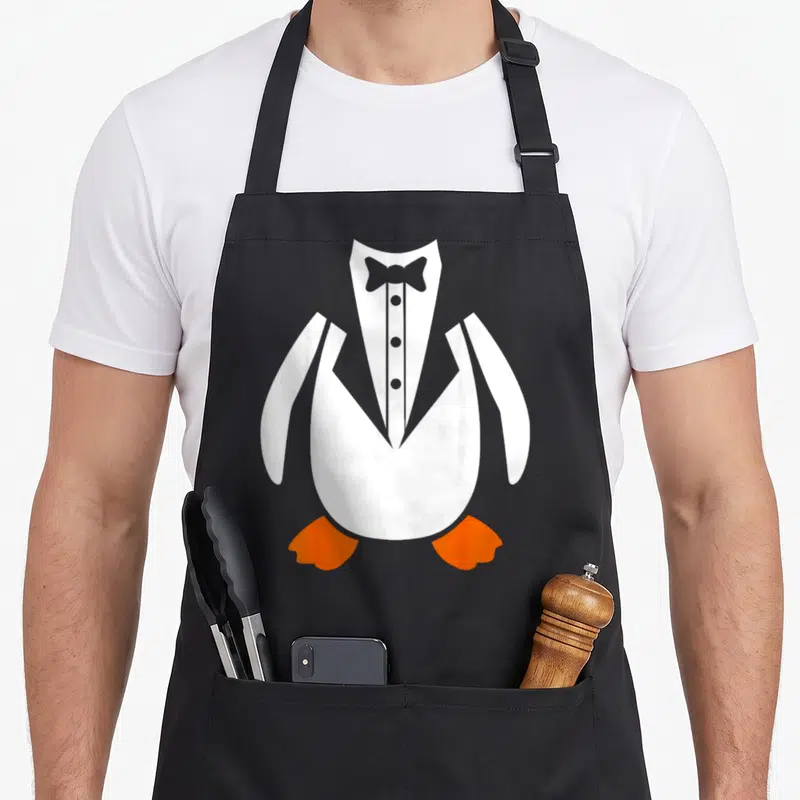 Tablier de cuisine noir avec illustration d'un pingouin vêtu d'un costume frac blanc et d'un noeud papillon gris.