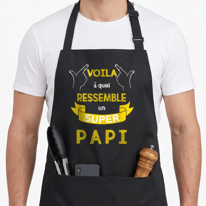 Tablier de cuisine noir avec une inscription jaune et blanche Voila à quoi ressemble un super papi encadrée par deux mains pointant vers le haut.