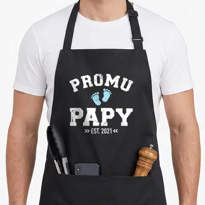Tablier de cuisine noir avec le texte promu papy est 2021 et deux empreintes de pieds bleues au centre.