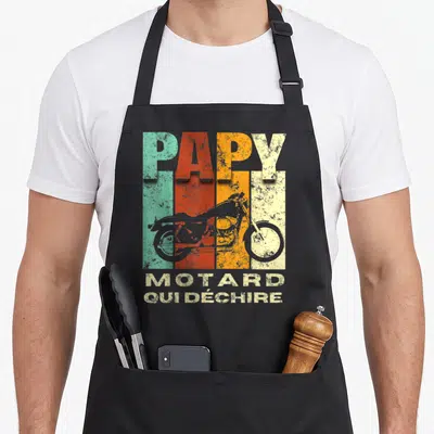 Tablier de cuisine noir avec linscription Papy Motard Qui Déchire et une illustration de moto vintage sur fond de bandes colorées.
