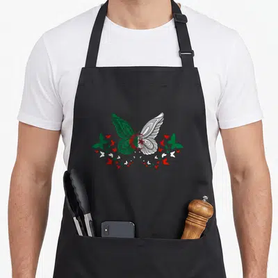 Tablier de cuisine noir avec grand papillon aux ailes vertes et blanches entouré de petits papillons rouges et blancs.