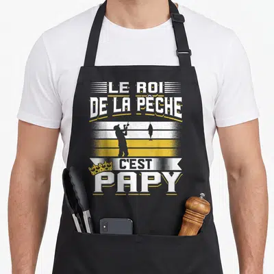 Tablier de cuisine noir avec le texte Le Roi de la Pêche C'est Papy et une silhouette de pêcheur tenant une canne.
