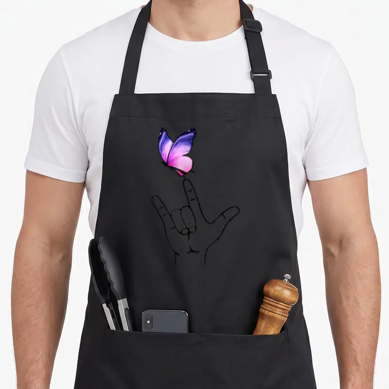 Tablier de cuisine noir avec une main en signe de love et un papillon violet et rose posé sur l'index.