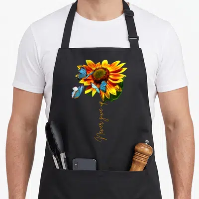 Tablier de cuisine noir avec une grande fleur de tournesol jaune et trois papillons bleus aux ailes déployées.