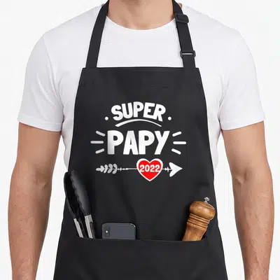 Tablier de cuisine noir avec inscription Super Papy 2022 et cœur rouge traversé par une flèche blanche stylisée.