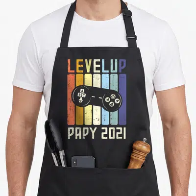 Tablier de cuisine noir avec illustration manette de jeu vidéo colorée et texte Level Up Papy 2021 en style vintage.
