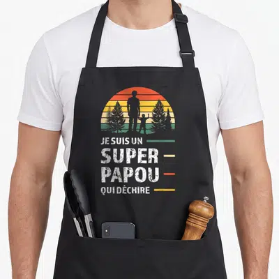 Tablier de cuisine noir avec silhouette d'homme et enfant devant un soleil couchant coloré et texte blanc.