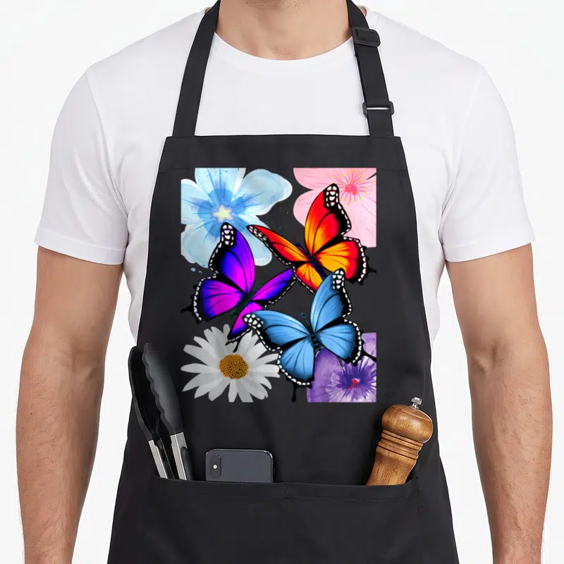 Tablier de cuisine noir avec des papillons rose bleu violet jaune et des fleurs blanches roses bleues et violettes imprimés au centre.