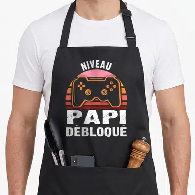 Tablier de cuisine noir avec illustration manette orange et texte Niveau Papi Débloqué en blanc.