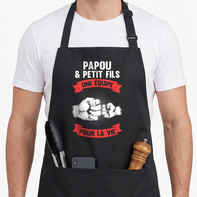 Tablier de cuisine noir avec illustration poings serrés et texte Papou et Petit Fils une équipe pour la vie sur banderoles rouges.
