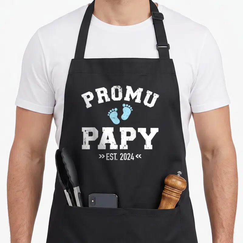 Tablier de cuisine noir avec Promu Papy Est. 2024 et traces de pas bleues en typographie blanche vintage.