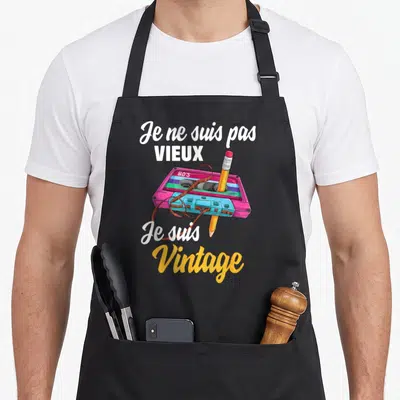 Tablier de cuisine noir avec une cassette rose et bleue et un crayon jaune traversant le texte blanc et orange.