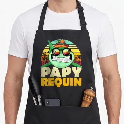 Tablier de cuisine noir avec un requin vert souriant portant un sombrero et des lunettes de soleil sur fond de coucher de soleil rétro.