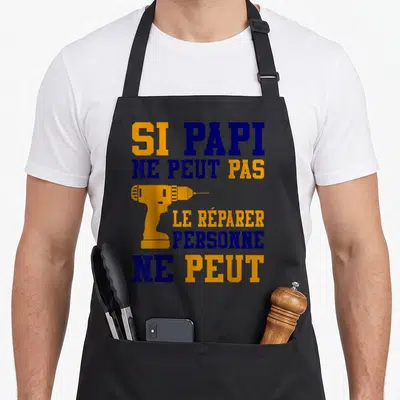 Tablier de cuisine noir avec texte SI PAPI NE PEUT PAS LE RÉPARER PERSONNE NE PEUT et une perceuse orange.