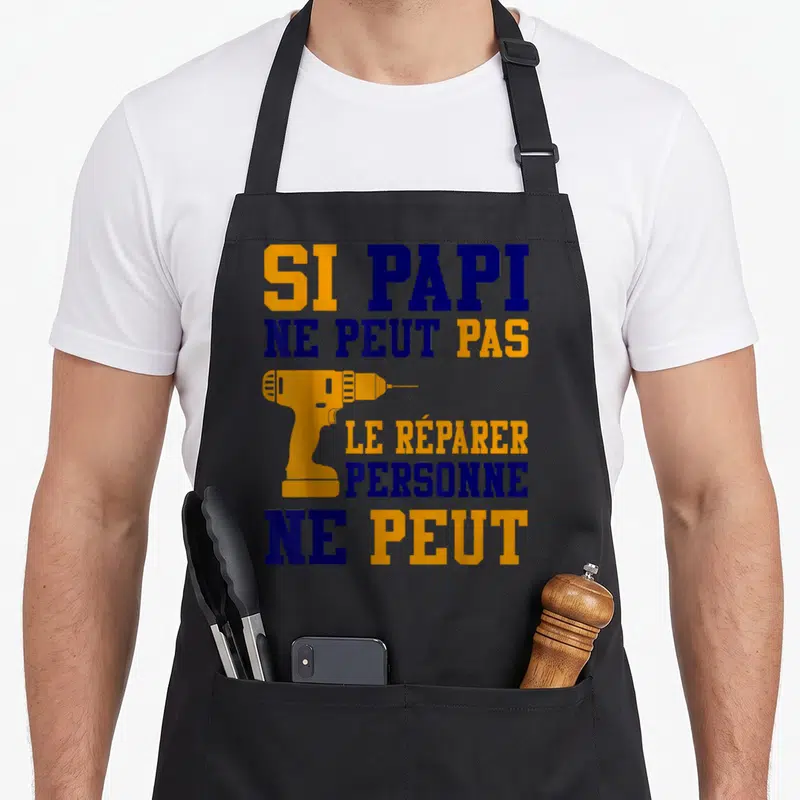 Tablier de cuisine noir avec texte SI PAPI NE PEUT PAS LE RÉPARER PERSONNE NE PEUT et une perceuse orange.