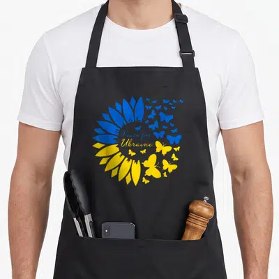 Tablier de cuisine noir avec un grand tournesol bleu et jaune formé de papillons et le texte Peace for Ukraine au centre.