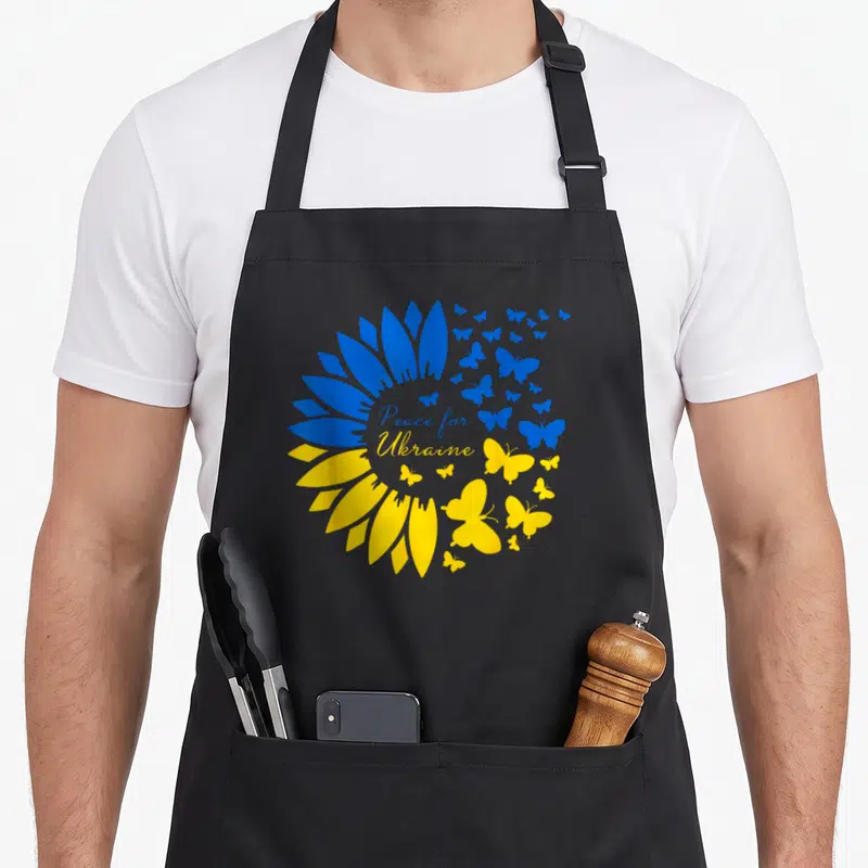 Tablier de cuisine noir avec un grand tournesol bleu et jaune formé de papillons et le texte Peace for Ukraine au centre.