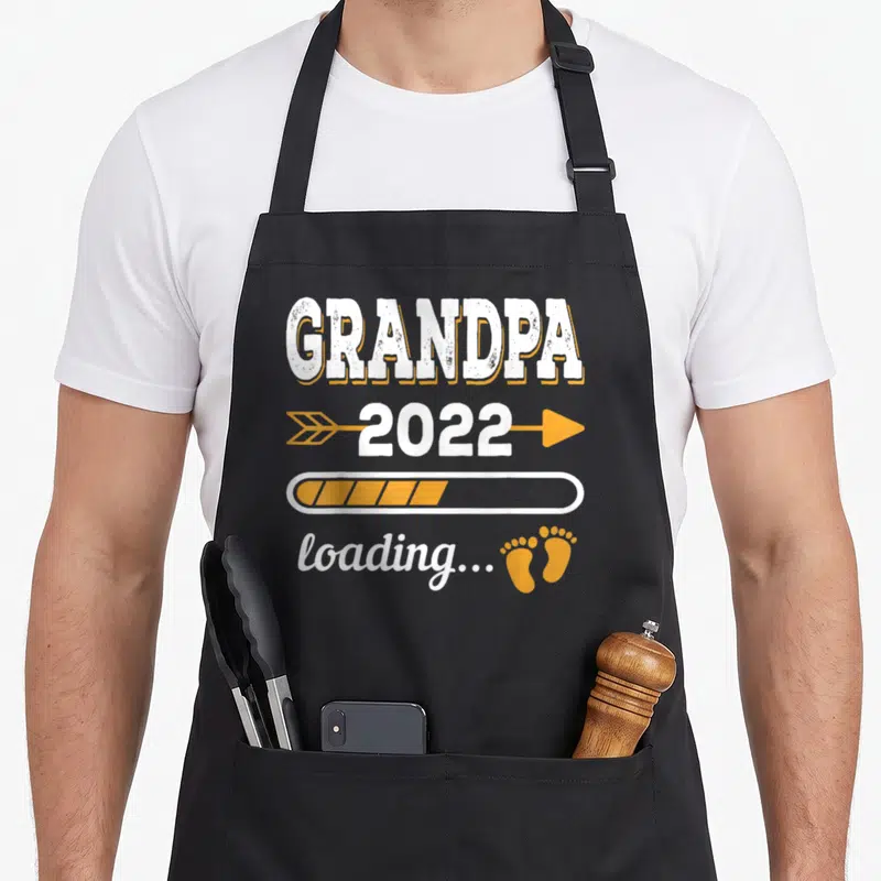 Tablier de cuisine noir avec l'inscription Grandpa 2022 loading et des petits pas orange.