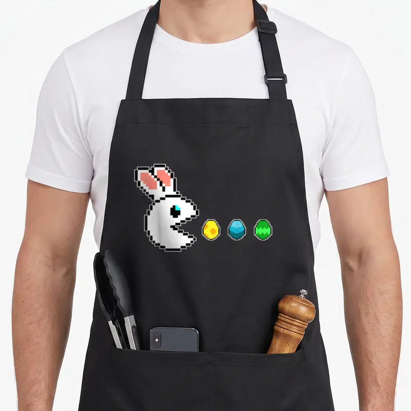 Tablier de cuisine noir avec lapin blanc pixel art type Pac-Man devant trois œufs de Pâques jaunes bleus et verts.