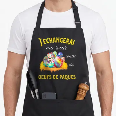Tablier de cuisine noir avec inscription jaune et illustration de lapin blanc parmi des œufs de Pâques multicolores sur une nasse dorée.