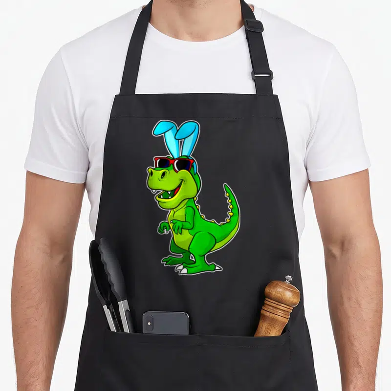 Tablier de cuisine noir avec un dinosaure vert portant des oreilles de lapin et des lunettes de soleil rouges.