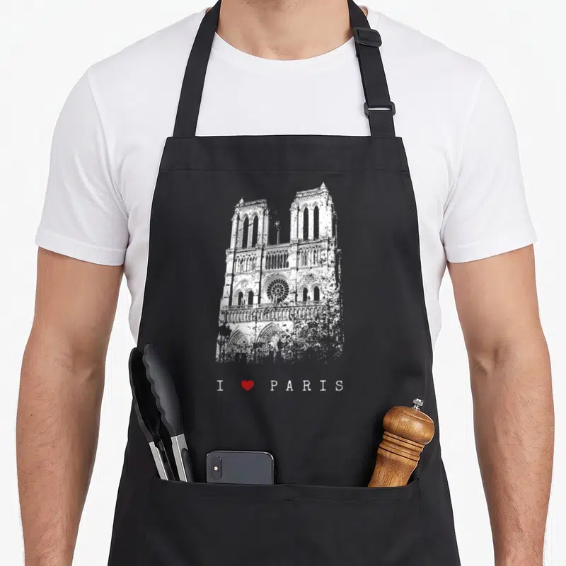 Tablier de cuisine noir avec illustration blanche de la cathédrale Notre-Dame et texte J'aime I Love Paris sur fond gris uni.