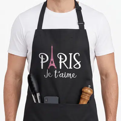 Tablier de cuisine noir avec l'inscription Paris Je t'aime et une Tour Eiffel rose stylisée au centre du motif imprimé.