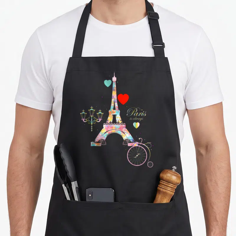 Tablier de cuisine noir avec une illustration colorée de la Tour Eiffel, des cœurs et un vélo rose stylisé sur fond uni.