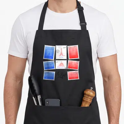 Tablier de cuisine noir avec stamps colorés représentant des monuments parisiens comme la Tour Eiffel et la cathédrale Notre-Dame.