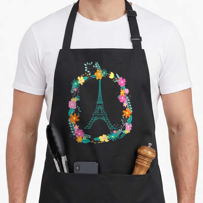 Tablier de cuisine noir avec une Tour Eiffel turquoise encadrée par une couronne de fleurs colorées et des feuilles stylisées.