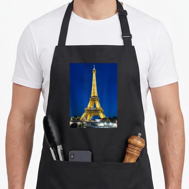 Tablier de cuisine noir avec une photo nocturne rehaussée de la Tour Eiffel à Paris illuminée sous un ciel bleu profond.