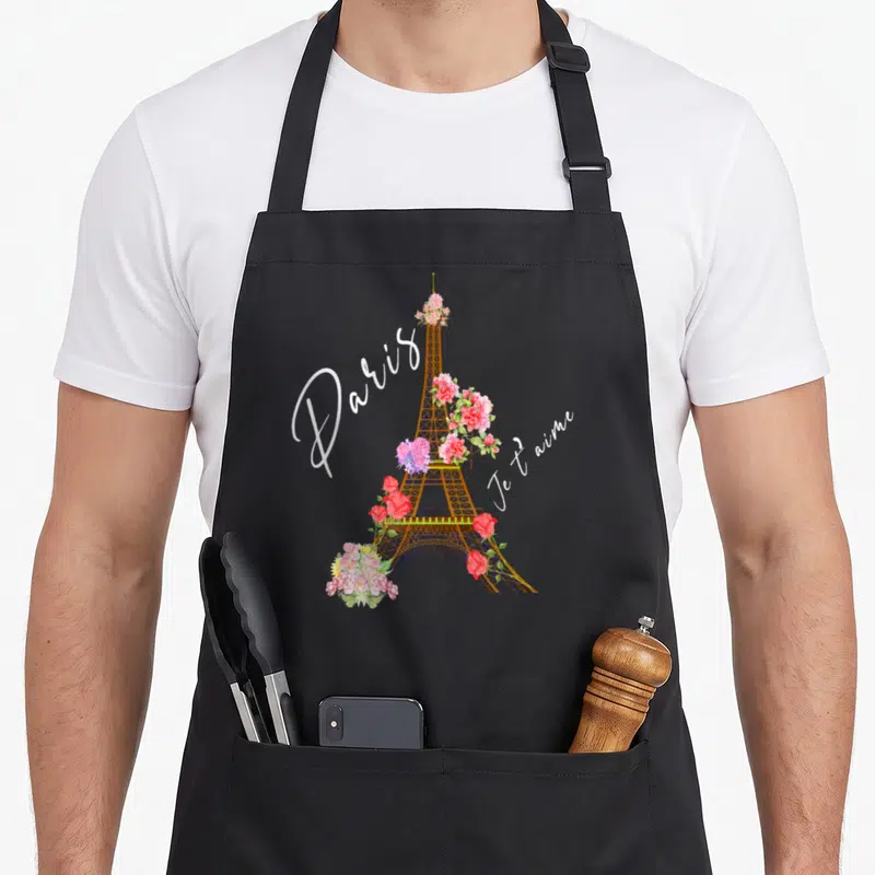 Tablier de cuisine noir avec une Tour Eiffel décorée de roses et d'hortensias, surmontée du texte Paris Je t'aime en calligraphie blanche.