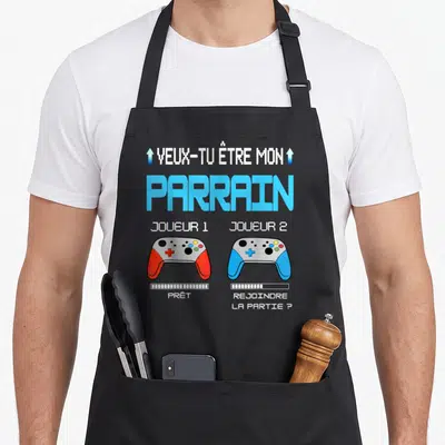 Tablier de cuisine noir avec deux manettes de jeu vidéo colorées et texte Veux Tu Être Mon Parrain en style pixel art.