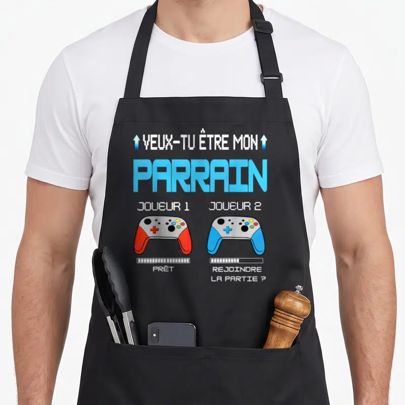 Tablier de cuisine noir avec deux manettes de jeu vidéo colorées et texte Veux Tu Être Mon Parrain en style pixel art.
