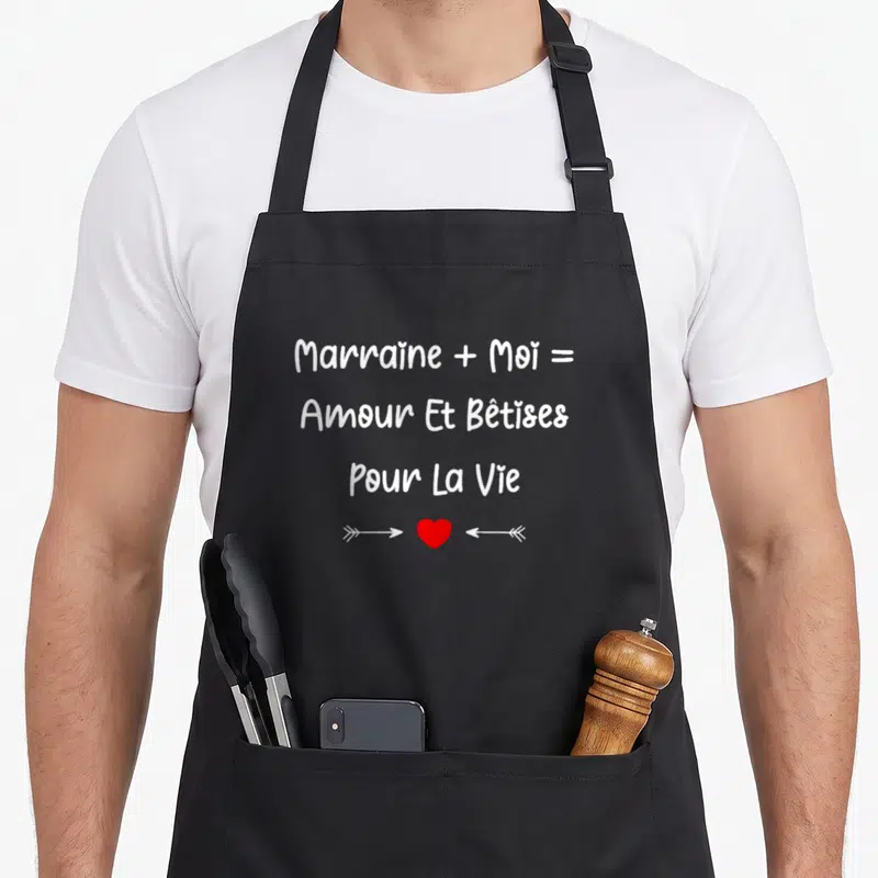 Tablier de cuisine noir avec l'inscription Marraine plus Moi égal Amour et bêtises pour la vie au-dessus d'un cœur rouge encadré par deux flèches blanches.