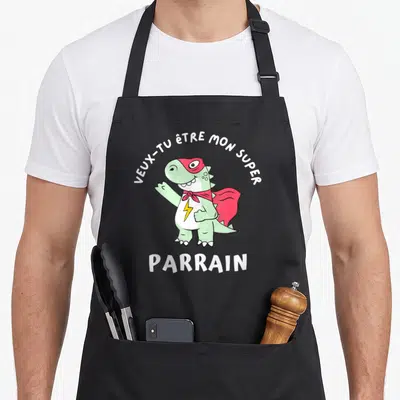 Tablier de cuisine noir avec un dinosaure super-héros vert portant un masque rouge et une cape, entouré du texte veux-tu être mon super parrain.