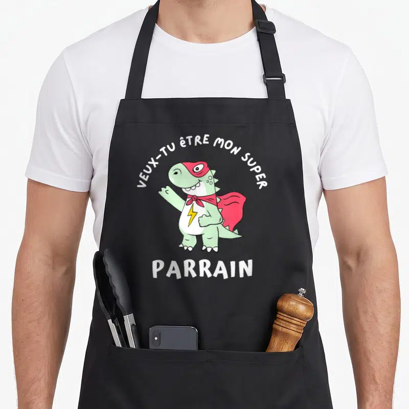Tablier de cuisine noir avec un dinosaure super-héros vert portant un masque rouge et une cape, entouré du texte veux-tu être mon super parrain.