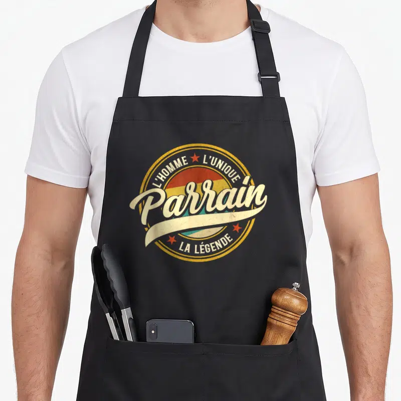 Tablier de cuisine noir avec logo circulaire vintage affichant Parrain L'homme l'unique la légende en lettres blanches et étoiles rouges.