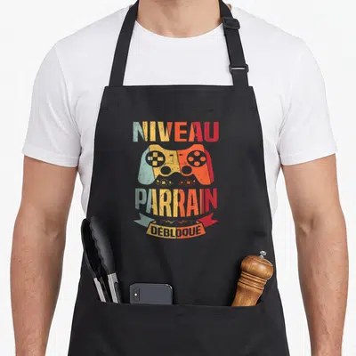 Tablier de cuisine noir avec inscription Niveau Parrain débloqué et illustration de manette de jeu vidéo multicolore style rétro.