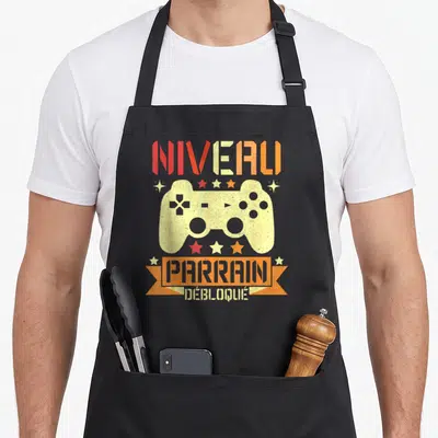 Tablier de cuisine noir avec illustration manette de jeu vidéo et texte Niveau Parrain Débloqué en couleurs chaudes.