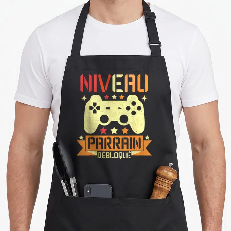 Tablier de cuisine noir avec illustration manette de jeu vidéo et texte Niveau Parrain Débloqué en couleurs chaudes.
