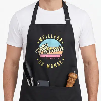 Tablier de cuisine noir avec motif circulaire retroMeilleur Parrain Du Monde sur fond de soleil pastel et silhouettes d'arbres.