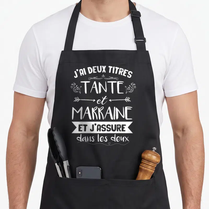 Tablier de cuisine noir avec l'inscription blanche Tante et Marraine et j'assure dans les deux entourée de motifs floraux et de flèches.