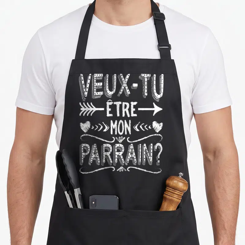 Tablier de cuisine noir avec l'inscription Veux Tu Etre Mon Parrain en blanc entouré de flèches et de cœurs dessinés.