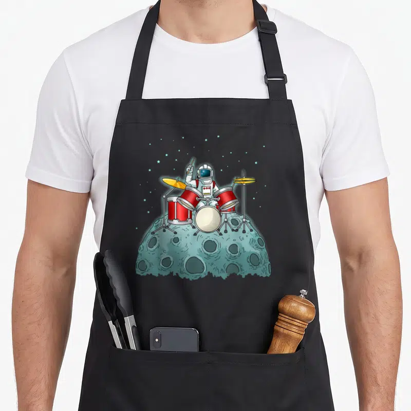 Tablier de cuisine noir avec illustration d'un astronaut jouant de la batterie sur un astéroïde lunaire étoilé.