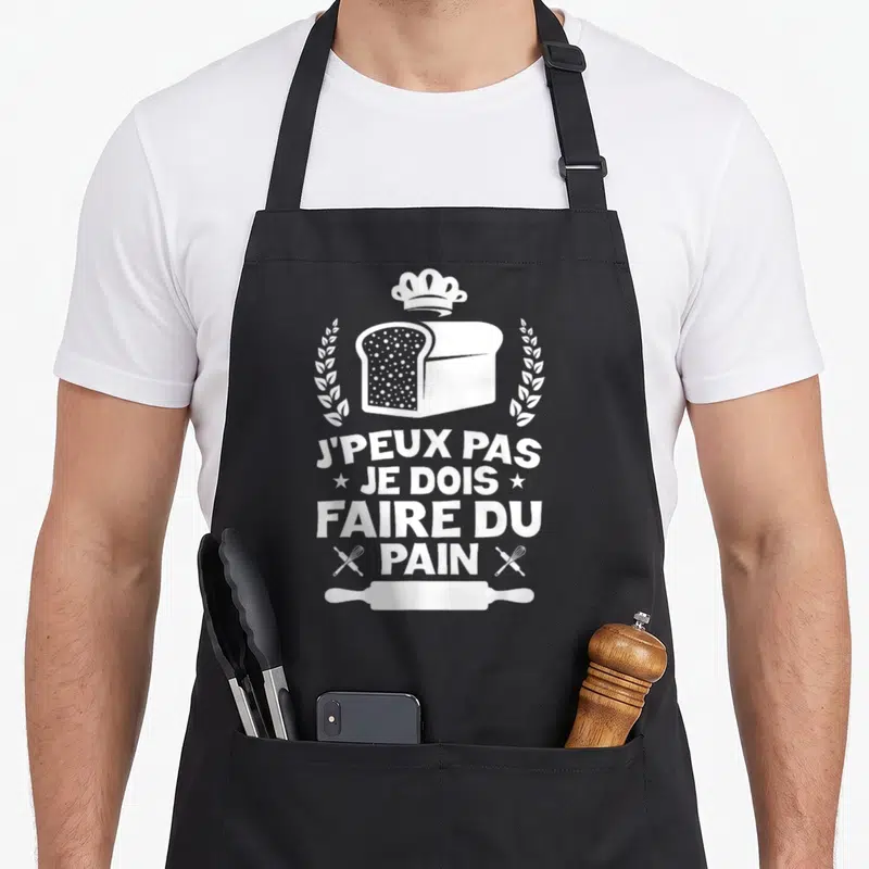 Tablier de cuisine noir avec illustration blanche d'un pain, chapeau de boulanger et texte humoristique.